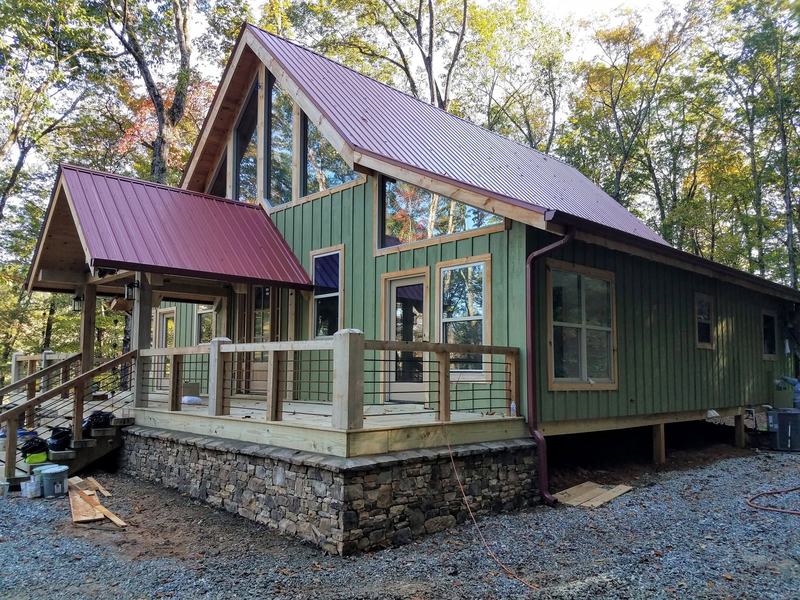 A green board-and-batten cabin with a metal roof, stone foundation and timber porch posts. Opens larger.