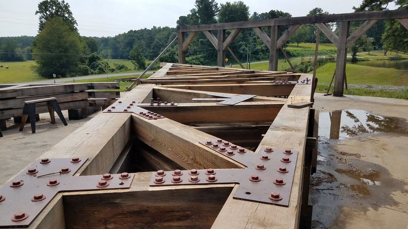 A timber truss laid out on the ground, its joints bolted with dark steel plates. Opens larger.